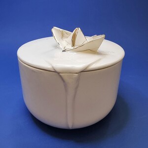 Può includere: Un contenitore in ceramica bianca con un coperchio. Il coperchio ha un design a forma di barca di carta dorata sulla parte superiore. Il contenitore ha una smaltatura bianca e una finitura liscia.
