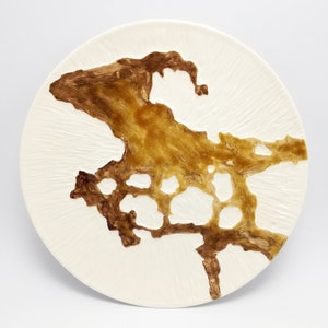 Può includere: Un piatto rotondo in ceramica bianco sporco con una superficie strutturata. Il piatto presenta un ampio design astratto nei toni del marrone e dell'oro, che ricorda una forma geografica. Il design ha un aspetto strutturato e stratificato, aggiungendo profondità.