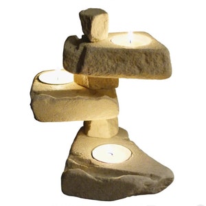 Puede incluir: Un portavelas de piedra apilada con tres velas de té. El soporte de color beige tiene un aspecto natural y texturizado. Las velas están encendidas, creando un brillo cálido.