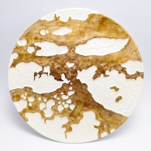 Può includere: Un piatto rotondo in ceramica bianco sporco con superficie strutturata. Il piatto presenta un motivo marrone organico che ricorda una mappa o un paesaggio. Il design è in rilievo, creando un effetto tattile tridimensionale. Adatto per scopi decorativi.