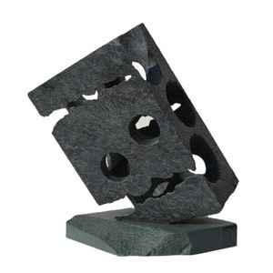 Puede incluir: Una escultura abstracta gris oscuro con forma de cubo, con cortes circulares y bordes irregulares. La escultura está montada sobre una base rectangular, creando una obra de arte moderna.