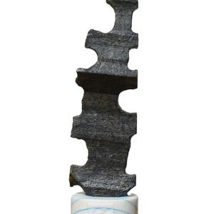 Puede incluir: Una escultura abstracta de color gris oscuro con una superficie rugosa y texturizada. La escultura tiene una forma irregular con secciones recortadas y se asienta sobre una base cilíndrica blanca y azul. El diseño general es moderno y artístico.