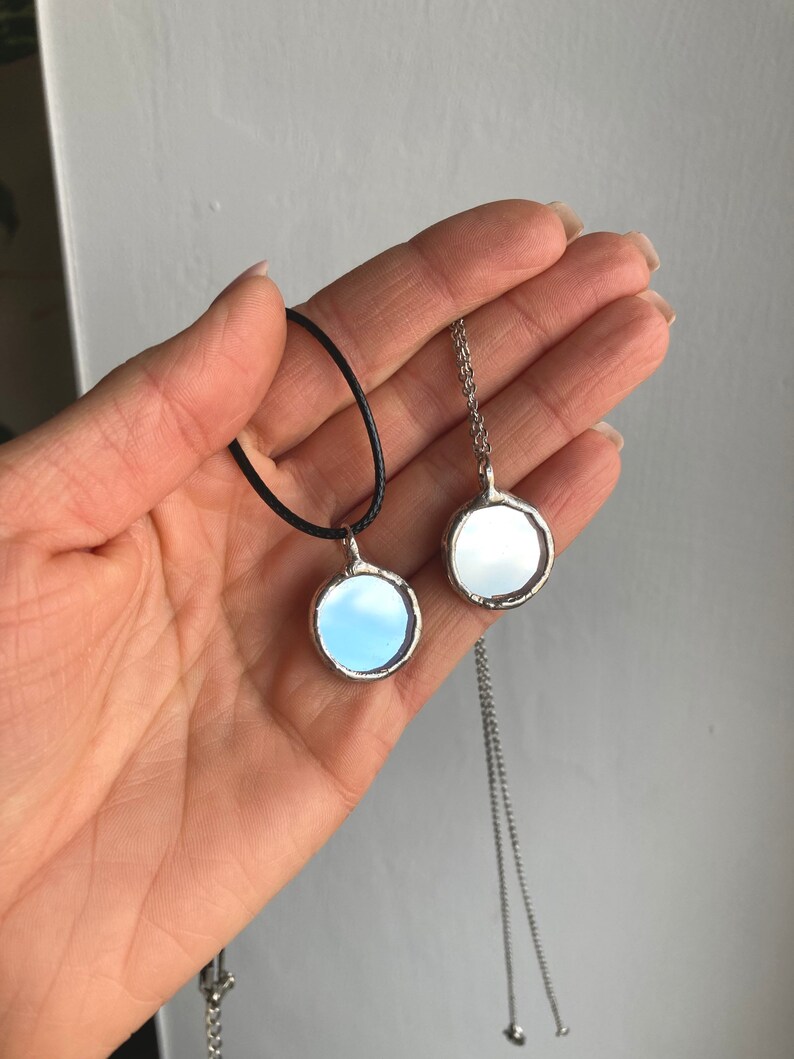 May include: Two silver necklaces with round mirror pendants. One necklace has a black cord and the other has a silver chain.