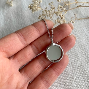 May include: A silver necklace with a round, textured pendant featuring a small, circular mirror.