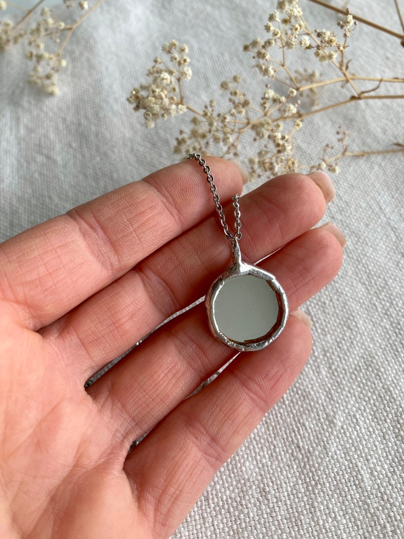 May include: A silver necklace with a round, textured pendant featuring a small, circular mirror.