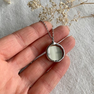 May include: A silver chain necklace with a round, silver pendant. The pendant has a smooth, white surface and a slightly irregular edge.