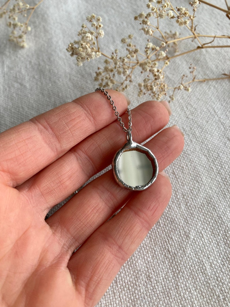 May include: A silver chain necklace with a round, silver pendant. The pendant has a smooth, white surface and a slightly irregular edge.