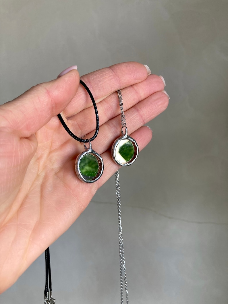 May include: Two silver necklaces with round green glass pendants. One necklace has a black cord and the other has a silver chain.