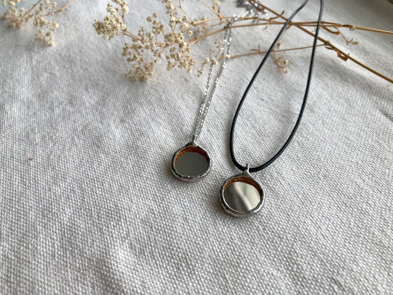May include: Two silver necklaces with round, mirrored pendants. One necklace has a black cord and the other has a silver chain.