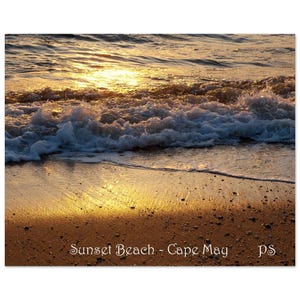 May include: A close-up of a sandy beach with foamy waves lapping at the shore. The water is reflecting the golden light of the setting sun, creating a shimmering effect on the sand. The text "Sunset Beach - Cape May" is visible in the lower right corner of the image.