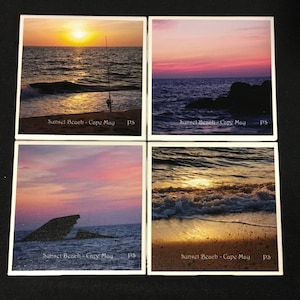 May include: Four square ceramic coasters with a sunset beach scene. Each coaster features a different sunset color palette with a rocky shoreline and ocean waves. The text "Sunset Beach - Cape May" is printed on each coaster.