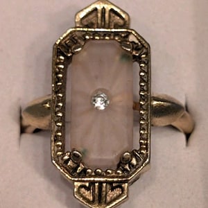 May include: A gold-toned ring featuring an elongated rectangular setting with a beaded border. The center contains a pale pink, faceted stone with a small, clear, round gemstone embedded in the middle. The metal frame has geometric cutouts.