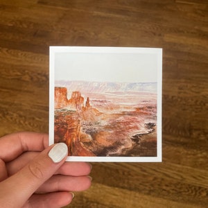 May include: A square print featuring a watercolor landscape of a canyon. The artwork depicts red rock formations, a vast canyon floor, and a hazy sky. The print is held by a hand against a wooden background.