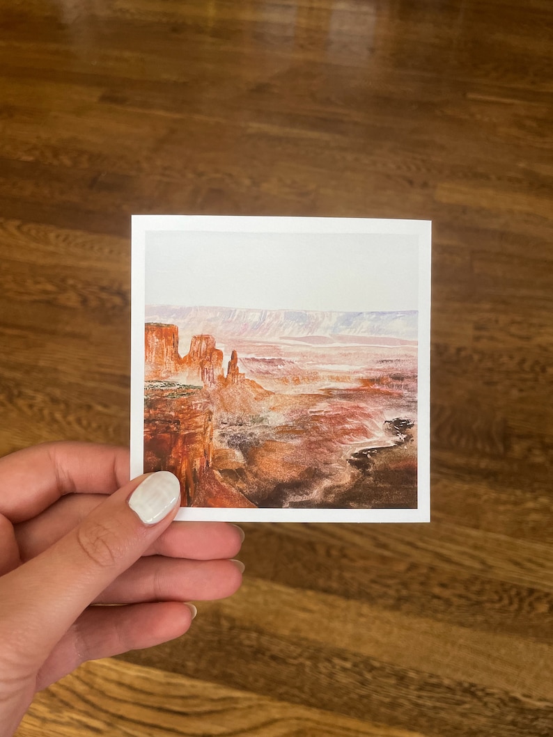 May include: A square print featuring a watercolor landscape of a canyon. The artwork depicts red rock formations, a vast canyon floor, and a hazy sky. The print is held by a hand against a wooden background.