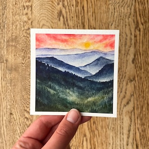 May include: Watercolor painting of a sunset over a mountain range. The sky is a vibrant red and orange, with a yellow sun peeking through the clouds. The mountains are a deep blue and green, with a forest in the foreground.