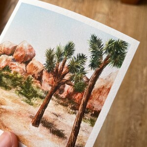 May include: Watercolor painting of a desert landscape featuring two Joshua trees with green foliage and brown trunks. The background includes reddish-brown rock formations and a light blue sky. The artwork is on textured paper.