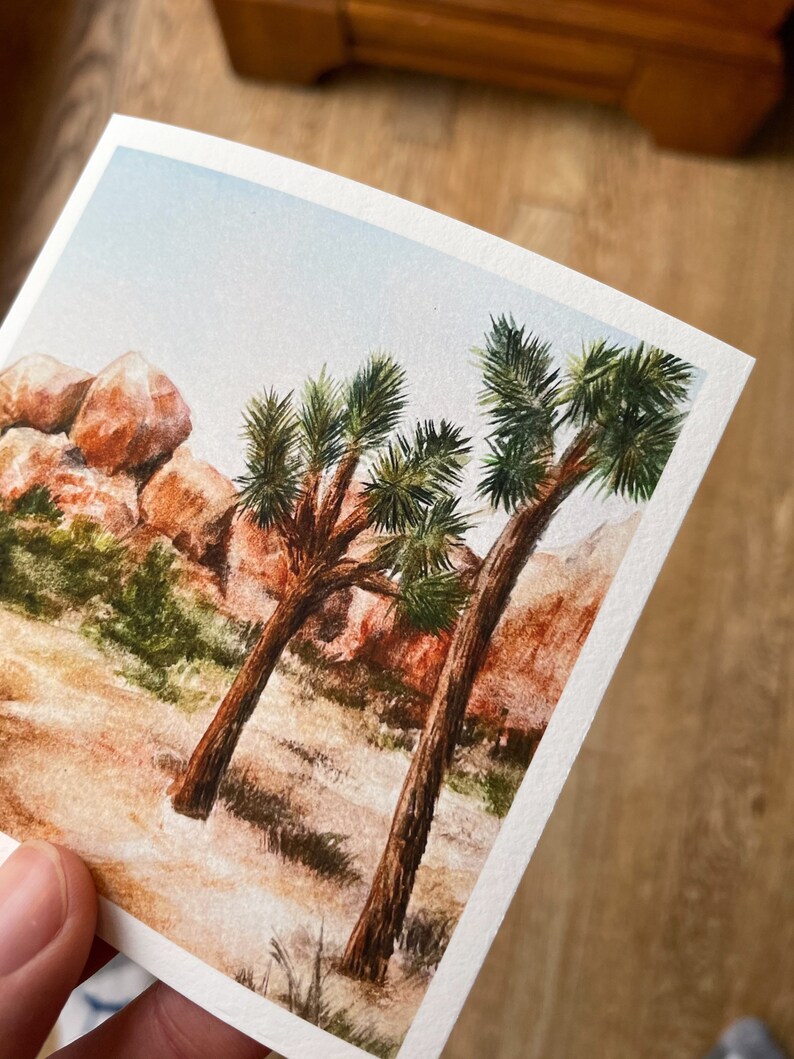 May include: Watercolor painting of a desert landscape featuring two Joshua trees with green foliage and brown trunks. The background includes reddish-brown rock formations and a light blue sky. The artwork is on textured paper.