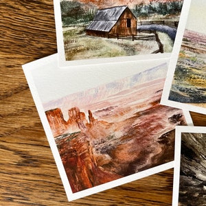 May include: A collection of watercolor landscape paintings featuring scenes of barns, mountains, and canyons. The paintings showcase a range of colors, including reds, browns, greens, and blues. Each painting is on white paper and is displayed on a wooden surface.