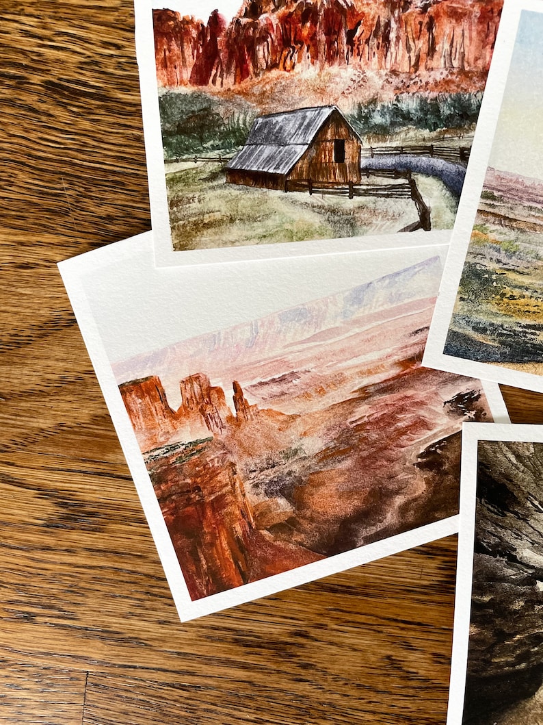 May include: A collection of watercolor landscape paintings featuring scenes of barns, mountains, and canyons. The paintings showcase a range of colors, including reds, browns, greens, and blues. Each painting is on white paper and is displayed on a wooden surface.