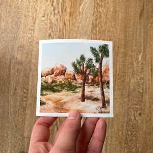 May include: A square watercolor print depicting a desert landscape with Joshua trees, rocks, and sparse vegetation. The artwork features warm tones of brown, beige, and green, with a light blue sky in the background. The print is held by a person.