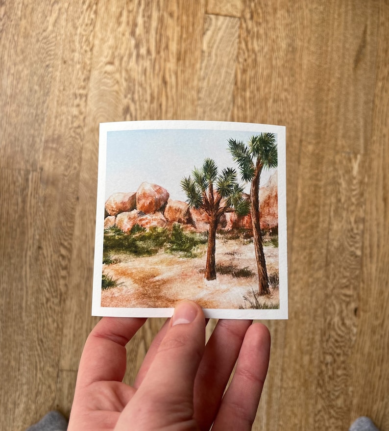 May include: A square watercolor print depicting a desert landscape with Joshua trees, rocks, and sparse vegetation. The artwork features warm tones of brown, beige, and green, with a light blue sky in the background. The print is held by a person.