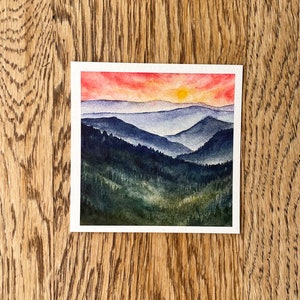 May include: Watercolor painting of a sunset over a mountain range. The sky is a vibrant red and orange, with a yellow sun peeking over the horizon. The mountains are a deep blue and green, with a hazy atmosphere.