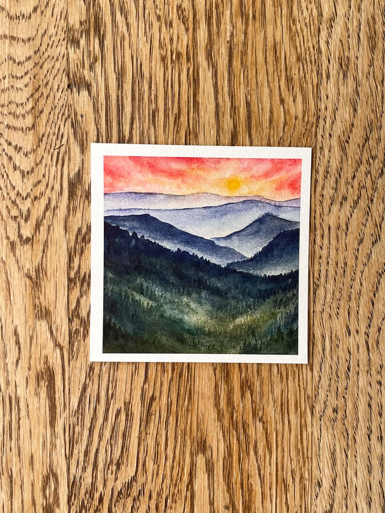 May include: Watercolor painting of a sunset over a mountain range. The sky is a vibrant red and orange, with a yellow sun peeking over the horizon. The mountains are a deep blue and green, with a hazy atmosphere.