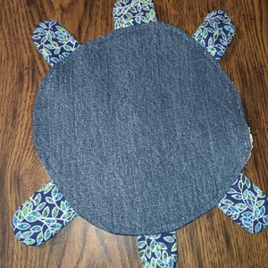Könnte beinhalten: Ein handgefertigtes, schildkrötenförmiges Objekt aus Jeansstoff. Der Körper der Schildkröte ist aus einfarbigem blauem Jeansstoff, mit gemusterten Flossenakzenten in blau, grün und weißen Blattdesigns. Auf einer Holzoberfläche platziert.
