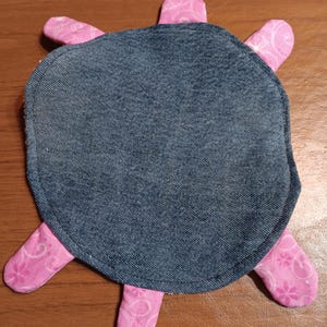 May include: A handmade turtle-shaped coaster. The coaster features a denim blue body and pink patterned legs. The pink fabric has a floral design. The coaster is designed to protect surfaces from heat and moisture.