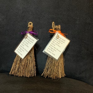 May include: Two miniature brooms with natural-colored bristles and twine handles. Each broom has a small tag with text, tied with a purple or orange ribbon. The text reads "Nothing Spills, Overcooks or Over Boils, This Kitchen Witch..." and continues.