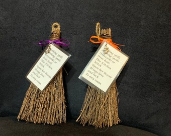 May include: Two miniature brooms with natural-coloured bristles and twine handles. Each broom has a small tag with text, tied with a purple or orange ribbon. The text reads "Nothing Spills, Overcooks or Over Boils, This Kitchen Witch..." and continues.