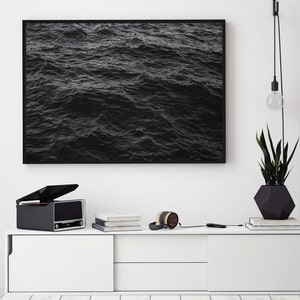 Peut inclure: Une impression encadrée de vagues sombres de l'océan est accrochée au-dessus d'un meuble blanc. Une platine vinyle noire, des livres et un casque sont posés sur le meuble. Un pot géométrique noir avec une plante et une suspension sont également présents.