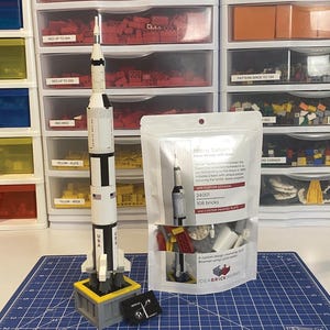 May include: A white, black, and gray model rocket with "USA" printed on the side, standing on a gray and yellow base. A clear bag with the text "Micro Saturn" and "108 bricks" is next to the rocket. The rocket is on a blue cutting mat.