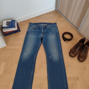 Op de afbeelding: Blauwe denim jeans met een knoopsluiting, plat op een houten vloer. Een bruine leren riem en versleten bruine leren laarzen liggen naast de jeans. Een stapel boeken is zichtbaar op de achtergrond.