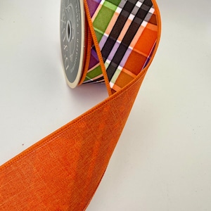 May include: A roll of decorative ribbon with a plaid pattern in orange, green, purple, black, and white. The ribbon has a solid orange edge and is likely used for crafts or gift wrapping.
