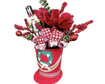 May include: A festive Christmas decoration featuring a red hat-shaped container filled with red flowers, berries, and a nutcracker figurine. The hat is adorned with a white and green wreath, gold trim, and patterned ribbons, creating a holiday centerpiece.