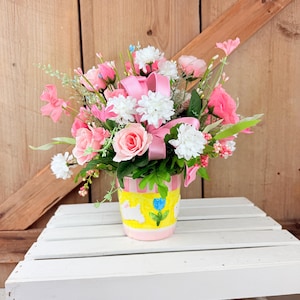 May include: A floral arrangement featuring pink roses, white chrysanthemums, and other flowers, accented with a pink ribbon. The flowers are arranged in a yellow and pink painted pot with a bunny and flower design. The arrangement is set against a wooden background.