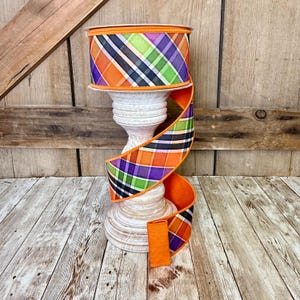 May include: A spool of decorative ribbon with a plaid pattern in orange, green, purple, and black. The ribbon is displayed on a white, carved pedestal against a rustic wooden background. The ribbon's edges are orange.