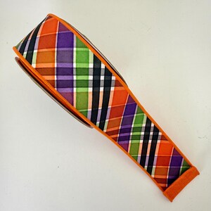 May include: A roll of decorative ribbon with a plaid pattern in orange, purple, green, and black. The ribbon has an orange border and is likely used for crafts or gift wrapping.