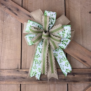 May include: A decorative bow featuring a mix of ribbons. The bow has a burlap base, with layers of white ribbon printed with green leaves, and green and beige lace trim. The bow is set against a rustic wooden background.