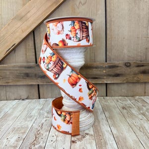 May include: A roll of decorative ribbon featuring a fall harvest theme. The white ribbon is printed with pumpkins, baskets, and autumn leaves in shades of orange, yellow, and green. The ribbon has a brown border.
