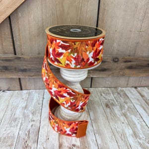 May include: A spool of decorative ribbon featuring a fall leaf pattern in shades of red, orange, and yellow on a white background. The ribbon is displayed on a white pedestal, with the spool's label visible. The ribbon is approximately 1.5 inches wide.