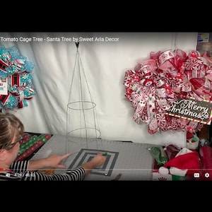 May include: A woman is making a Christmas tree out of a tomato cage. She is using a red and white wreath with a "Merry Christmas" sign as inspiration.