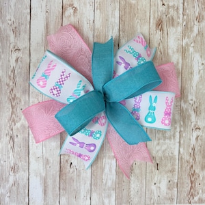May include: An Easter-themed bow with a turquoise centre and pink and white ribbons. The white ribbons feature patterned bunnies in purple, teal, and pink. The pink ribbons have a floral design. Ideal for Easter decorations.