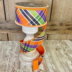 May include: A spool of decorative ribbon with a plaid pattern in orange, purple, green, and black. The ribbon is displayed on a white pedestal, with a length of ribbon cascading down. The background is a wooden surface.
