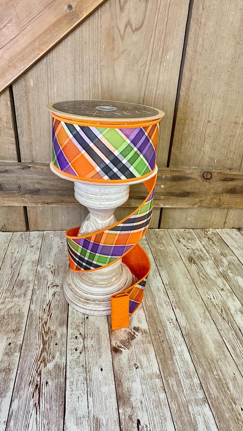 May include: A spool of decorative ribbon with a plaid pattern in orange, purple, green, and black. The ribbon is displayed on a white pedestal, with a length of ribbon cascading down. The background is a wooden surface.