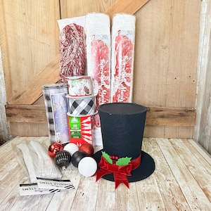 May include: A craft supply kit featuring a black top hat with a red velvet ribbon and holly, two rolls of white snowball mesh, a roll of black and white buffalo check ribbon, a roll of deco poly mesh, white chenille stems, and assorted ornaments.