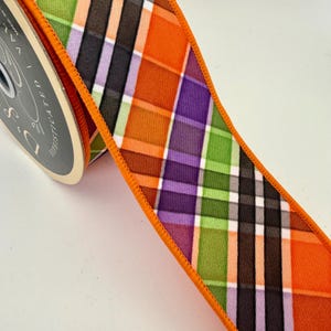 May include: A spool of decorative ribbon with a plaid pattern in orange, green, purple, black, and white. The ribbon has an orange stitched edge. The spool is labeled "D's Sophisticated".