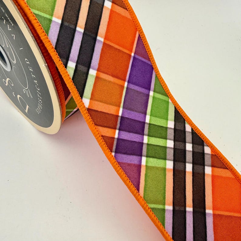 May include: A spool of decorative ribbon with a plaid pattern in orange, green, purple, black, and white. The ribbon has an orange stitched edge. The spool is labeled "D's Sophisticated".