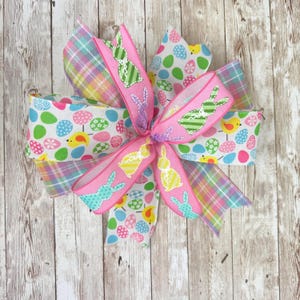 May include: An Easter-themed bow with pink, patterned, and plaid ribbons. The bow features a central pink ribbon with bunny designs. Other ribbons display Easter eggs, chicks, and pastel plaid patterns. The bow is set against a wooden background.